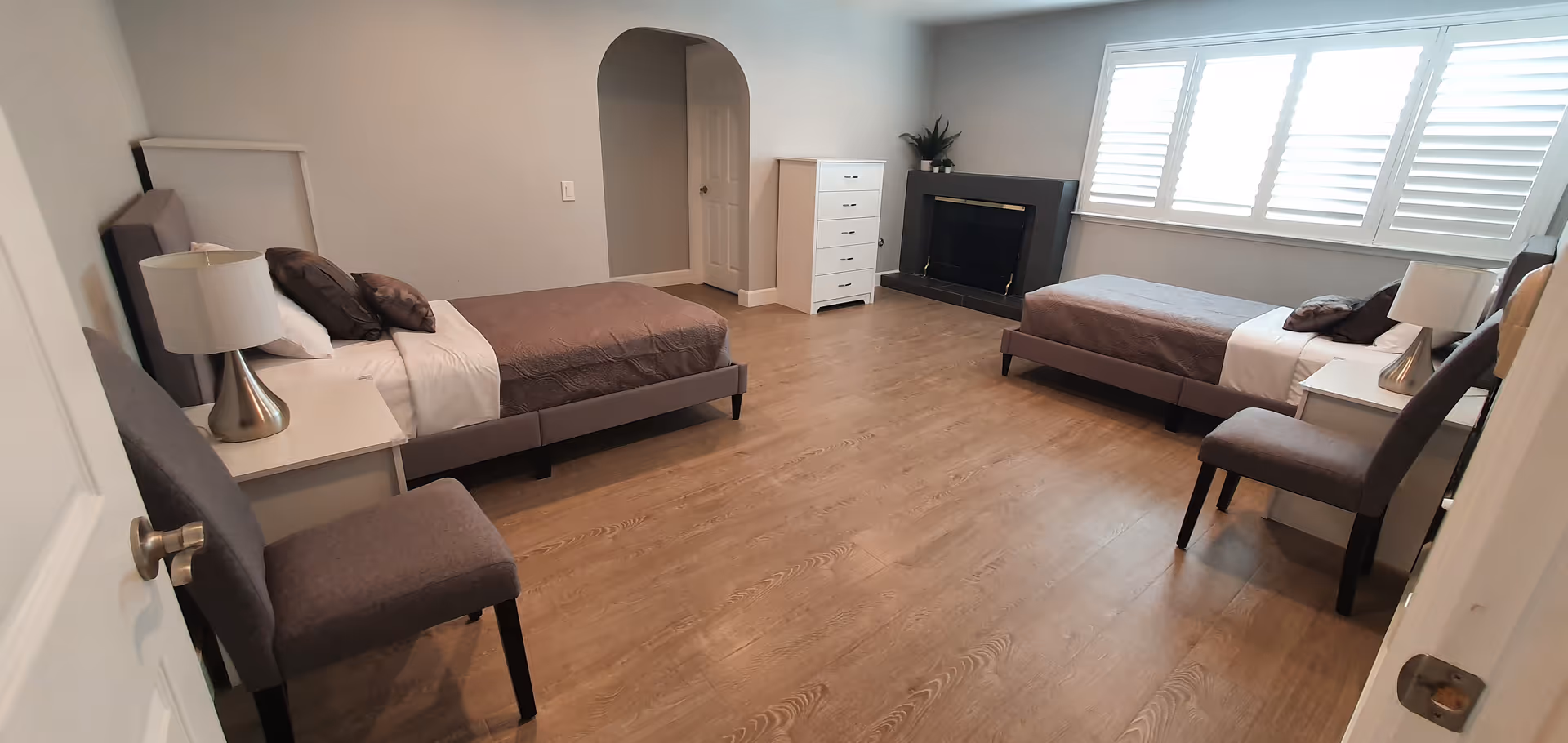 A spacious bedroom with two single beds, each with brown bedding and white pillows. There are two bedside tables with lamps, two chairs, a white dresser, a fireplace, and a large window with white shutters letting in natural light. The floor is wooden and the walls are painted light gray.