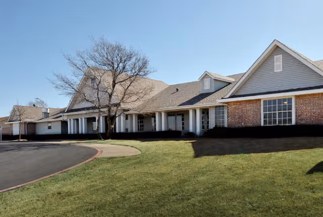 Exterior view of a single-story senior living facility building with a combination of brick and siding walls, multiple windows, a covered entrance, and a leafless tree on a grassy lawn under a clear blue sky.