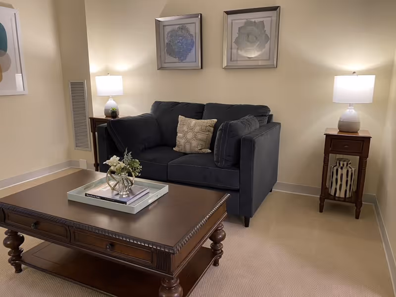 A cozy living room with a dark gray loveseat adorned with a patterned cushion, flanked by two wooden side tables each holding a white lamp. Above the loveseat are two framed floral artworks. In front of the loveseat is a large wooden coffee table with a decorative tray holding books and a small flower arrangement. The room has beige walls and carpeted flooring.