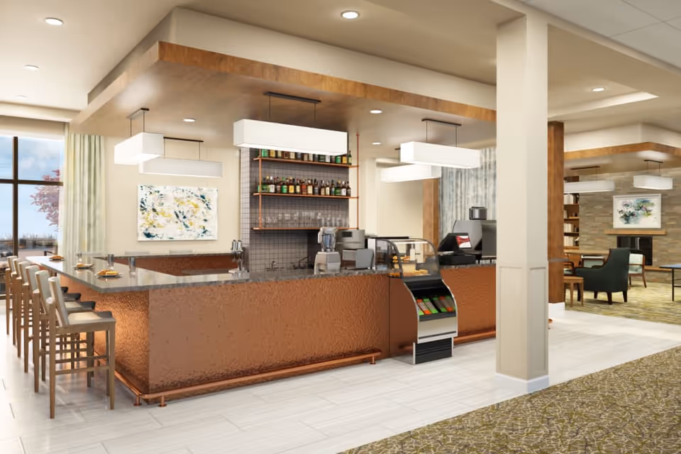 Interior view of a modern cafe or bar area with a long counter, bar stools, shelves with bottles, and pendant lighting. Adjacent seating area with chairs and tables is visible, along with large windows letting in natural light.