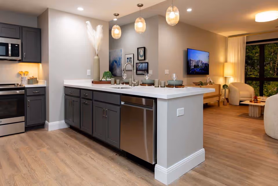 Modern kitchen area with dark gray cabinets, stainless steel appliances including a microwave, oven, and dishwasher, and a white countertop island with a sink and decorative items. The kitchen opens into a living room with a wall-mounted TV, a lamp, two armchairs, a coffee table, and large windows showing greenery outside.