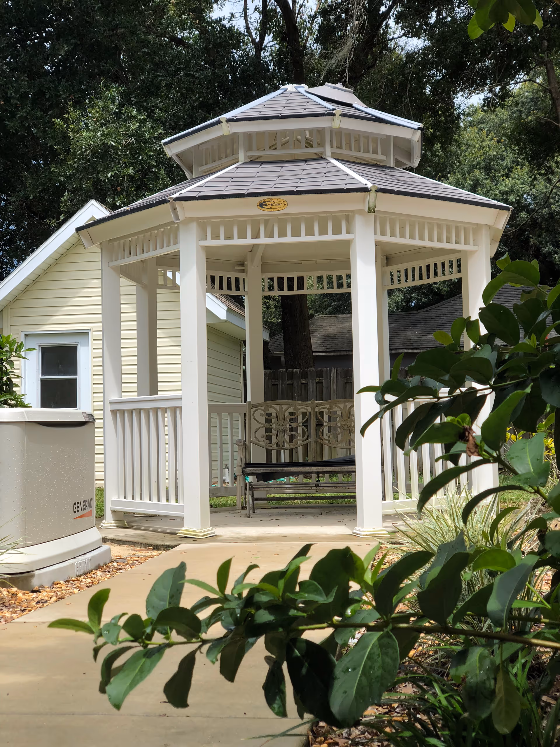 A white wooden gazebo with a shingled roof situated on a concrete pathway surrounded by greenery. There is a decorative metal bench inside the gazebo. In the background, there is a yellow building with white trim and a window. Trees and plants surround the area, creating a peaceful outdoor setting.