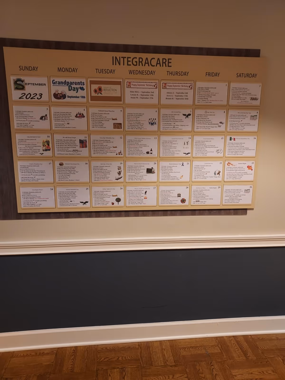 A bulletin board mounted on a beige wall displaying a monthly calendar for September 2023 titled INTEGRACARE, with daily activities and events listed for each day of the week. The calendar includes special days such as Grandparents Day and various scheduled activities. The wall below the bulletin board is painted dark blue with white trim, and the floor is wooden.