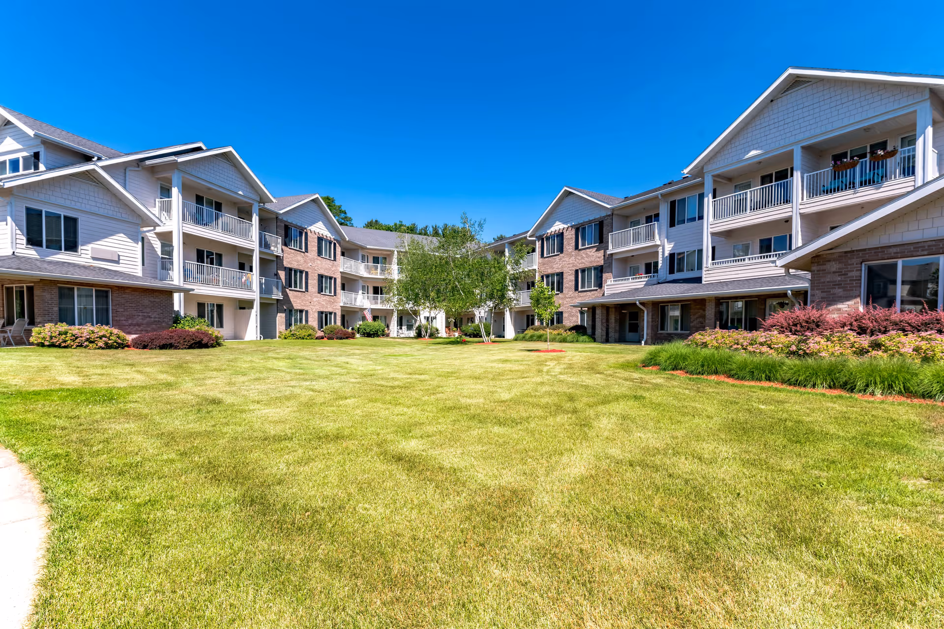 Three-story senior living complex with balconies surrounding a large green courtyard under a clear blue sky.