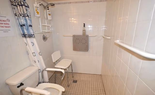 Accessible tiled bathroom with a shower area, shower chair, grab bars, and a toilet with armrests.