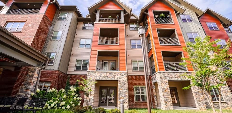Exterior view of a multi-story residential building with a combination of brick, stone, and siding in beige and orange tones. The building features balconies with railings, windows, and a landscaped area with green plants and white flowers in front. There is a lamppost near the entrance and outdoor seating visible on the left side.