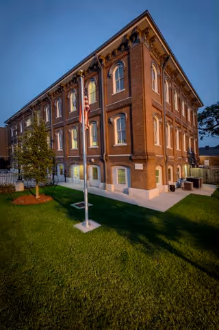 A three-story brick building with multiple arched windows, illuminated from inside during dusk. In front of the building is a well-maintained lawn with a flagpole displaying the American flag and a small tree. The sky is clear and the scene is peaceful.