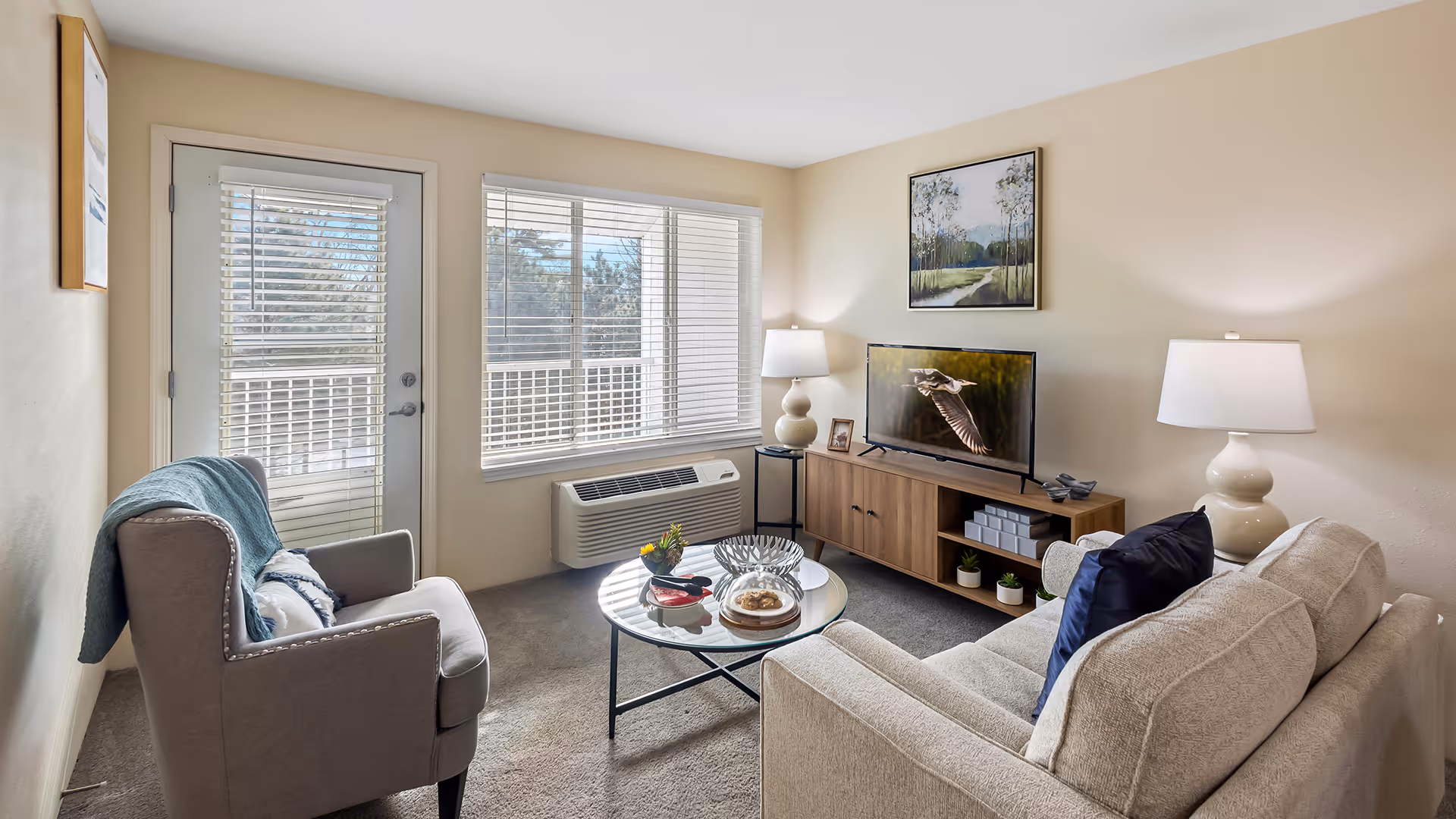 A cozy living room with a beige sofa and a gray armchair, both with cushions. A round glass coffee table is in the center with decorative items on it. There is a wooden TV stand with a flat-screen TV displaying a bird in flight. Two table lamps with white shades are on either side of the TV stand. A large window with white blinds and a glass door letting in natural light are visible. The walls are painted light beige and there is a framed landscape painting above the TV.