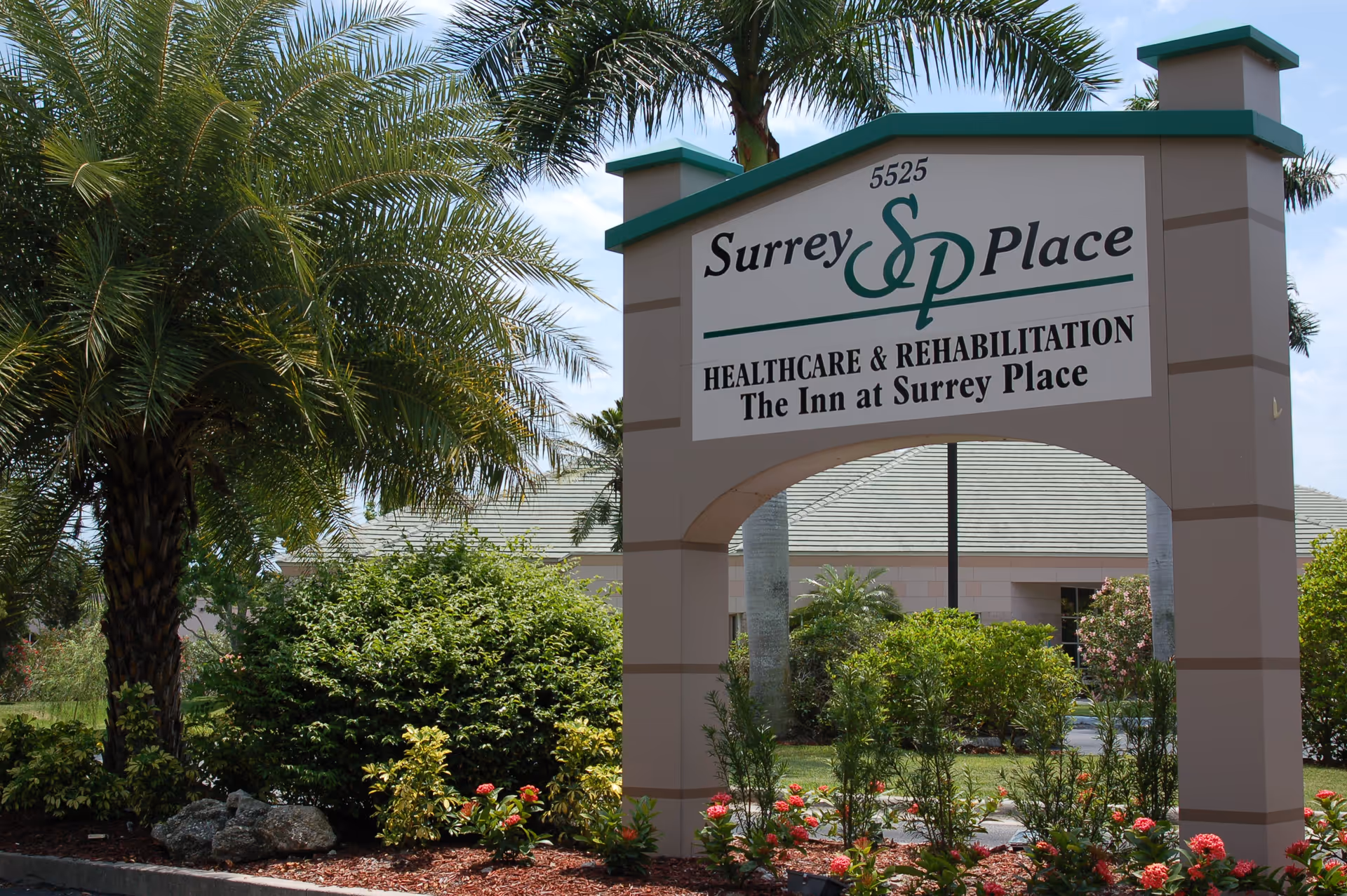 Outdoor view of the entrance sign for Surrey Place Healthcare & Rehabilitation, surrounded by palm trees, bushes, and flowering plants with a building visible in the background.