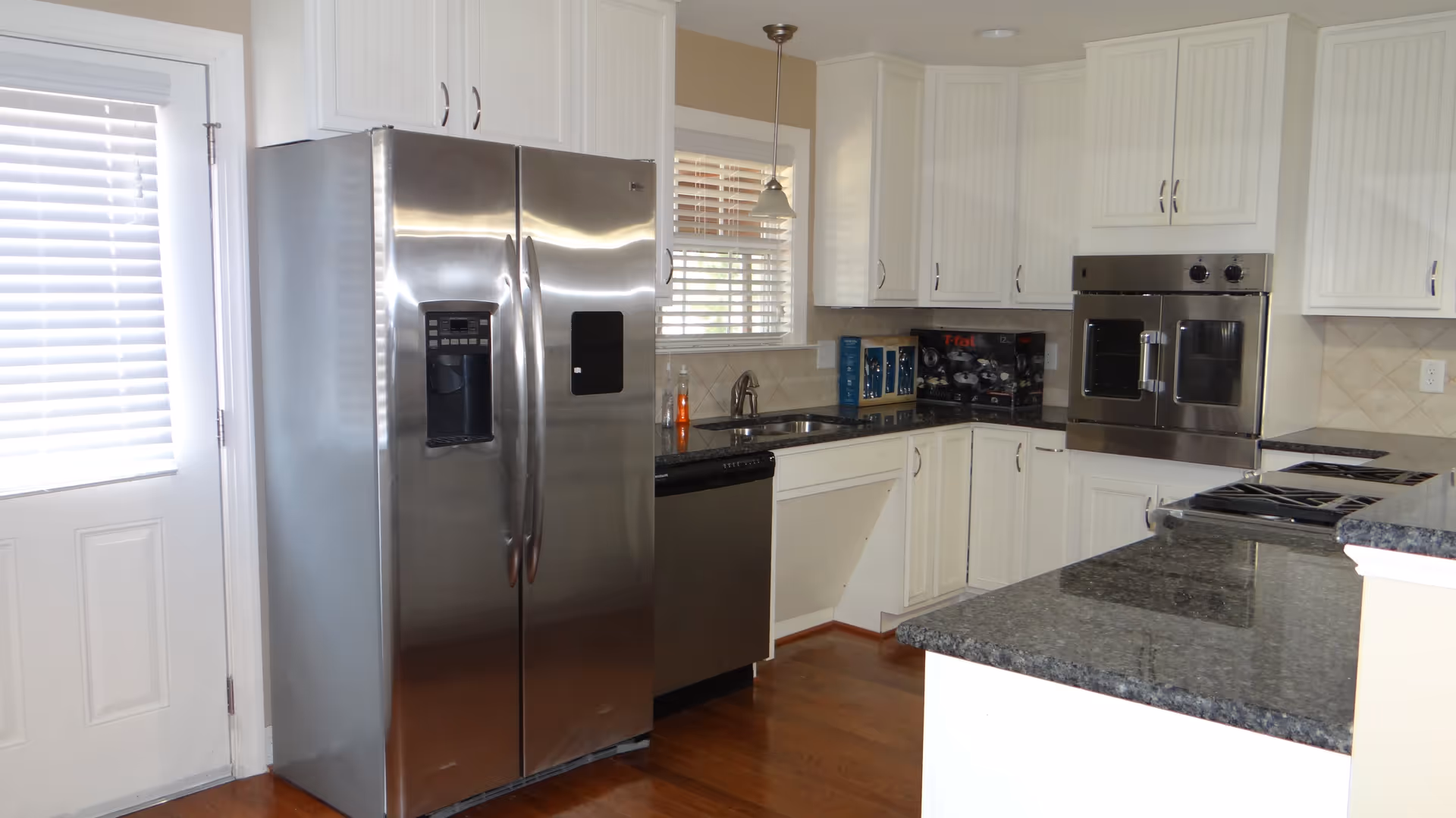 Bright modern kitchen with stainless steel side-by-side refrigerator, double oven, granite countertops, white cabinets and hardwood floors.