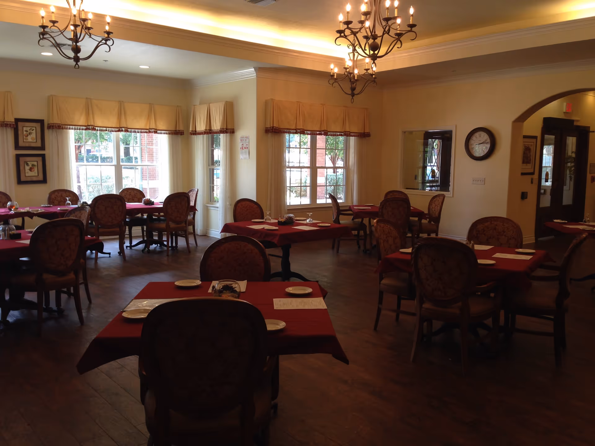 Dining room with multiple round tables covered in red tablecloths and upholstered chairs under chandeliers and large windows.