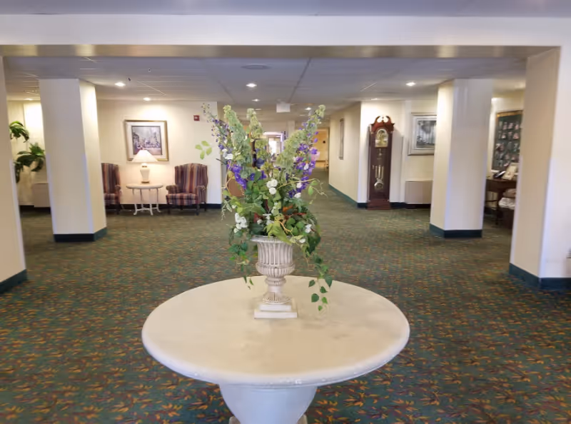 Interior view of a senior living facility hallway with a round table in the foreground holding a large floral arrangement. The hallway has patterned carpet, cream-colored walls, and ceiling tiles. There are chairs, a side table with a lamp, framed pictures on the walls, and a grandfather clock visible in the background.