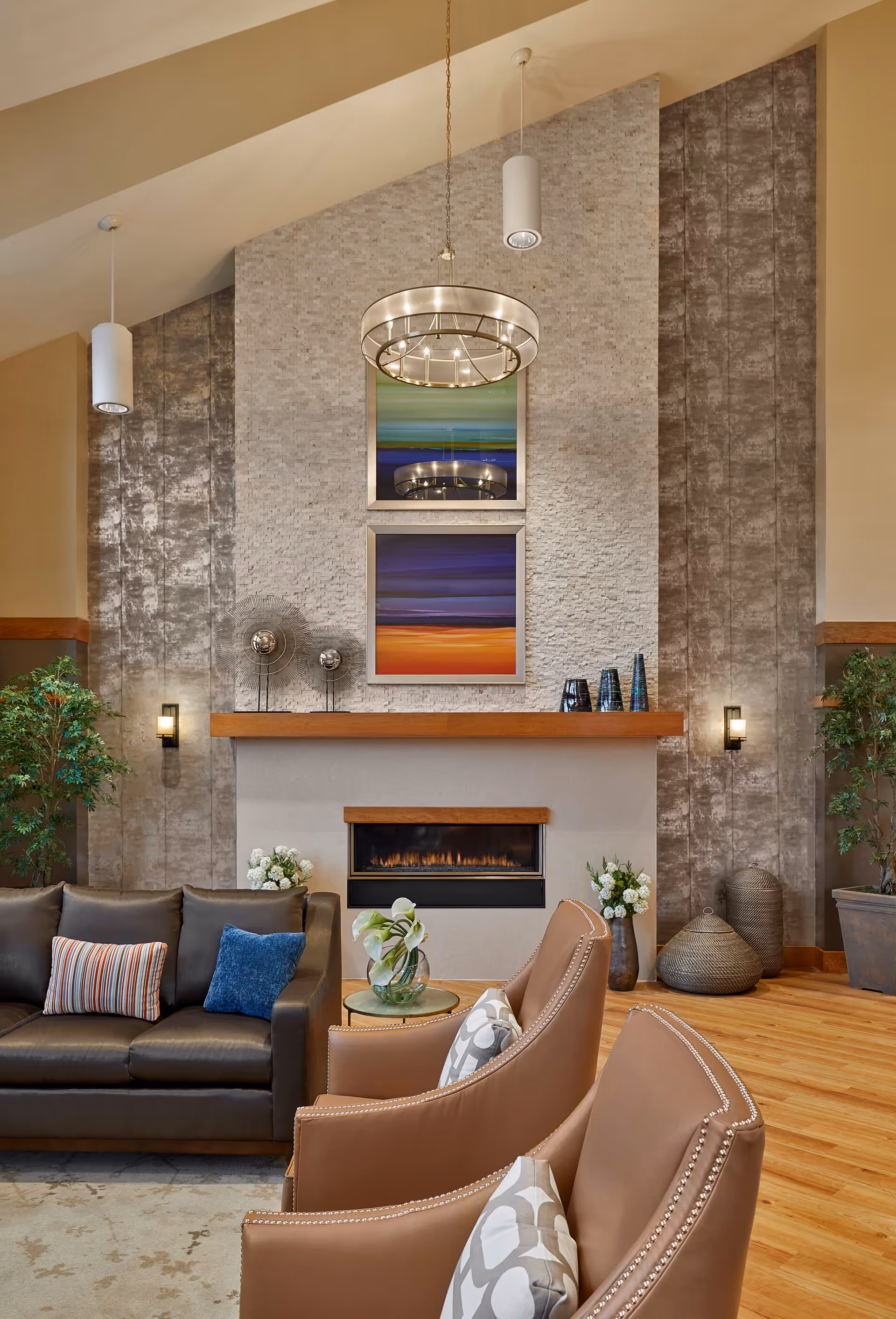 A cozy living room area in Leawood nursing home featuring a modern fireplace with a wooden mantel, two abstract colorful paintings above it, a dark leather sofa with striped and blue cushions, two tan leather armchairs with patterned pillows, a small round glass table with a vase of white flowers, decorative plants, and warm wooden flooring under soft lighting.