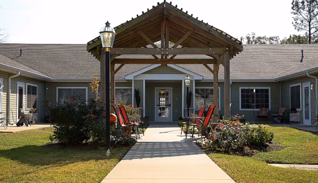 Outdoor courtyard area of Brookfield Assisted Living featuring a wooden pergola with a pathway leading to the building entrance. There are red cushioned chairs arranged on either side of the pathway, surrounded by green grass and flower bushes. The building has light green siding and multiple windows.