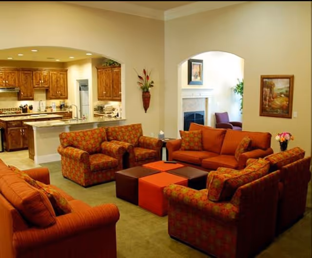 A cozy living room with orange patterned sofas and armchairs arranged around square ottomans, open to a kitchen with wooden cabinets.