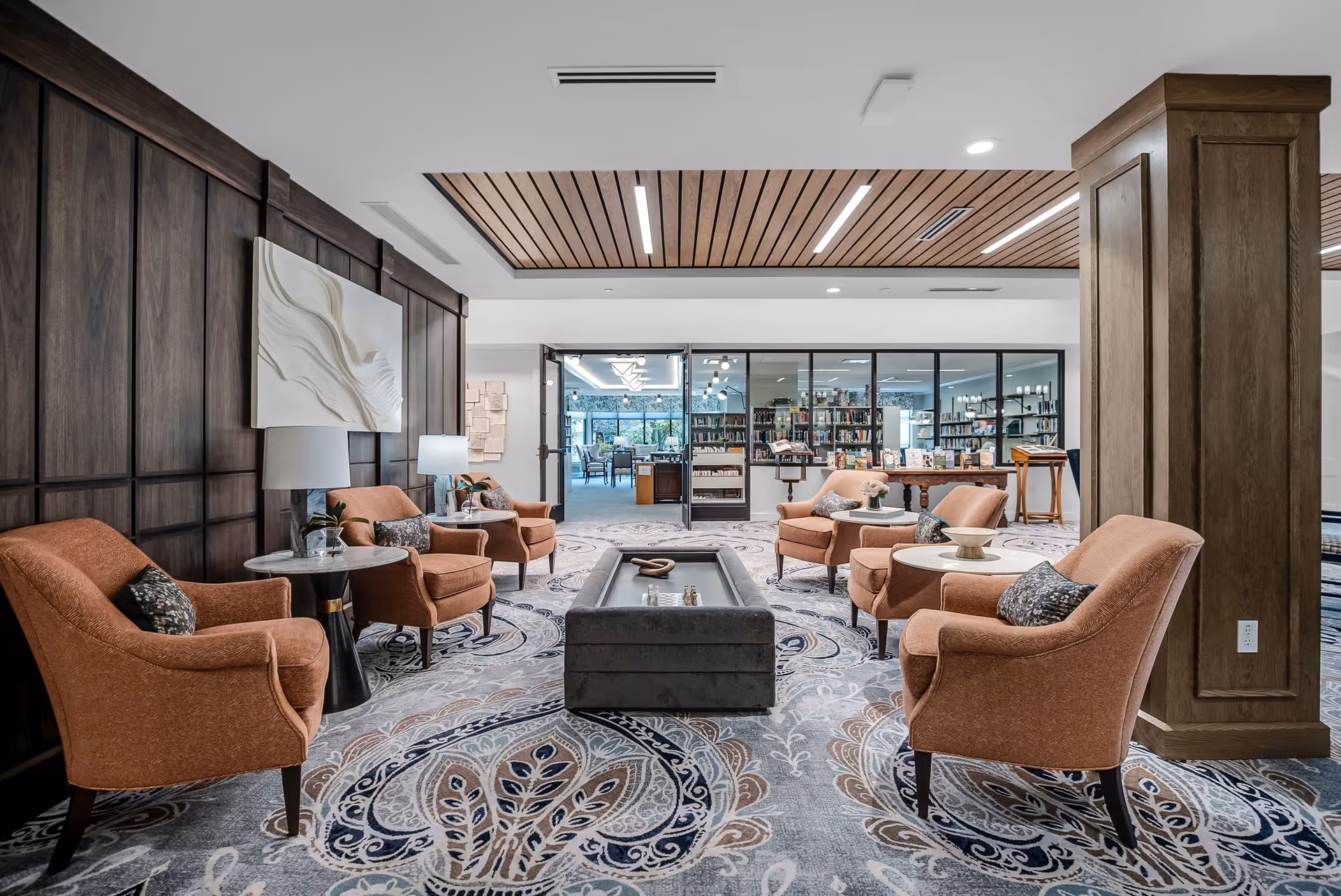 A bright communal lounge with upholstered armchairs and side tables arranged around a central ottoman, leading to a glass-fronted library.