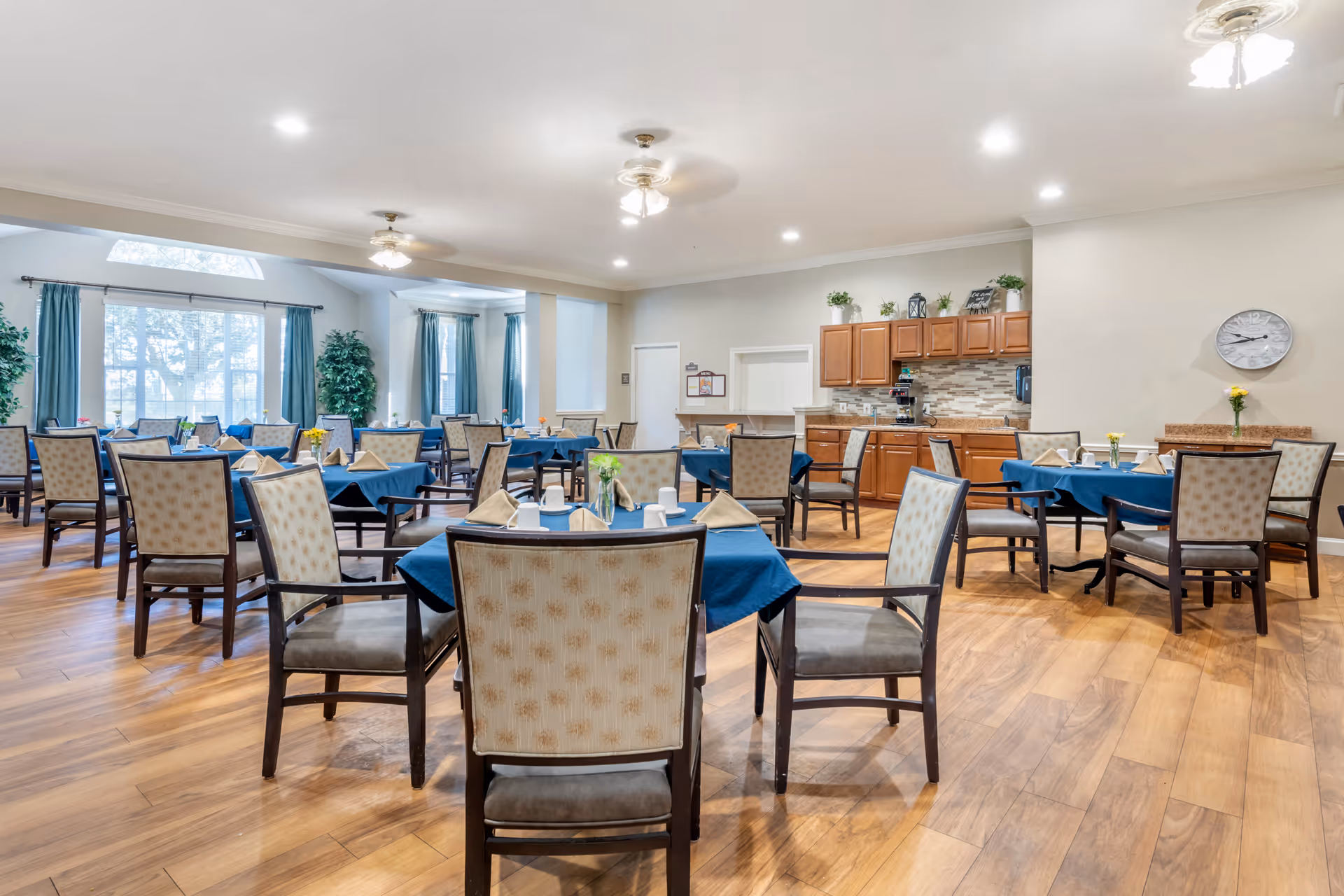 A bright dining room with multiple tables covered in blue tablecloths, each set with beige napkins, white cups, and small flower vases. The room has wooden flooring, beige walls, and large windows with teal curtains letting in natural light. There are ceiling fans and recessed lighting overhead, and a kitchen area with wooden cabinets and a coffee machine is visible in the background.