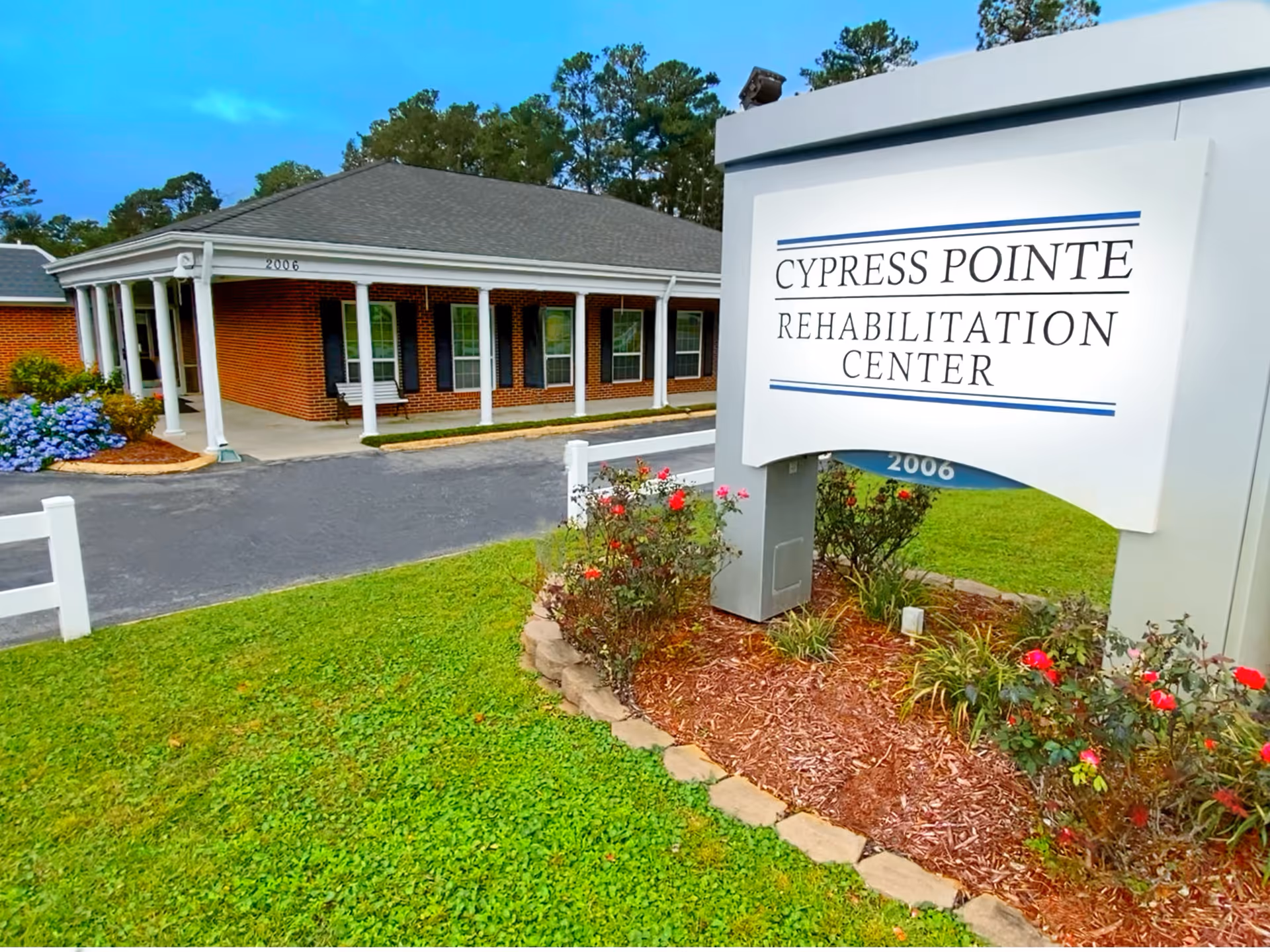 Exterior view of Cypress Pointe Rehabilitation Center showing a brick building with white columns and a covered porch. There is a large white sign with the facility's name in front, surrounded by a landscaped area with flowers and green grass.