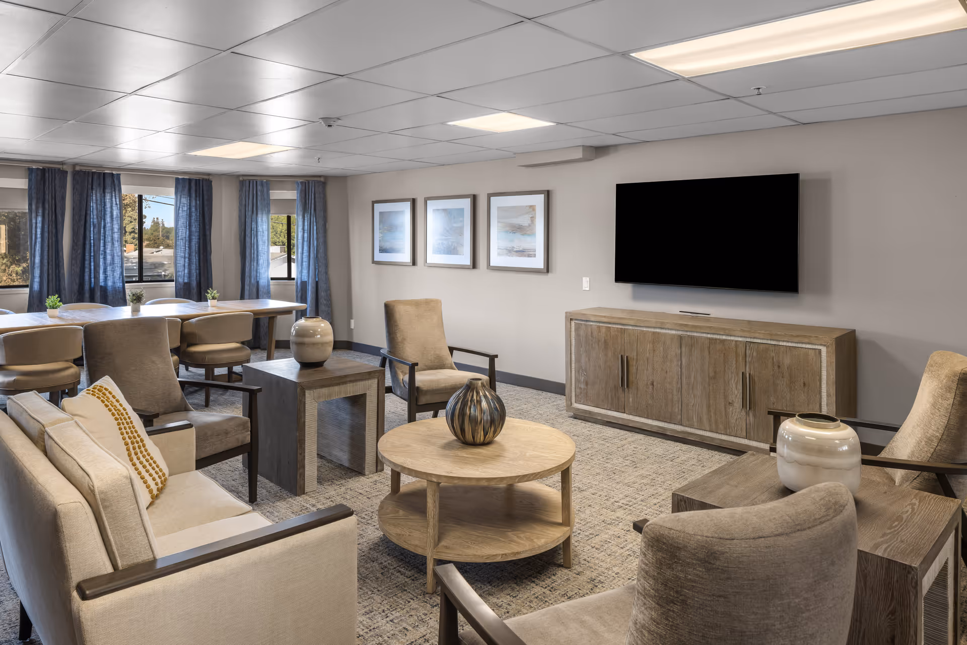 A cozy senior living community lounge with beige armchairs and a sofa arranged around a round wooden coffee table and side tables. The room features a large flat-screen TV mounted on the wall above a wooden cabinet, three framed artworks on the wall, and windows with blue curtains letting in natural light.