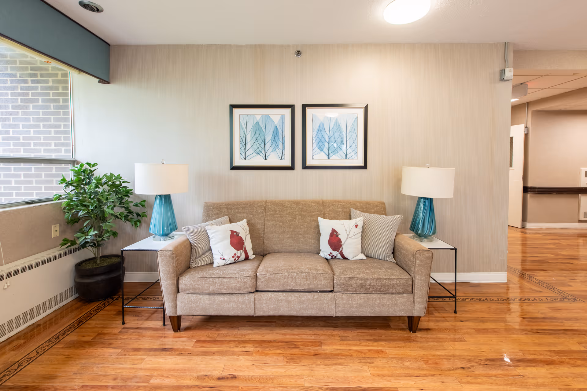 A beige sofa with four cushions, two featuring red cardinals and two plain, is centered against a light-colored wall. Above the sofa are two framed pictures of blue leaves. On either side of the sofa are small tables with blue glass lamps. To the left, there is a large potted green plant near a window. The floor is wooden with a decorative border.
