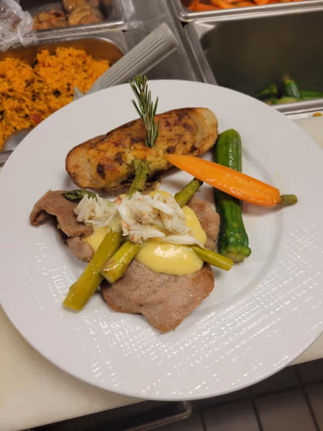 A plated meal consisting of two slices of cooked meat topped with asparagus and a creamy yellow sauce, garnished with shredded crab meat. The plate also includes a stuffed baked potato garnished with a sprig of rosemary, a cooked baby carrot, and a small zucchini. In the background, there are metal food trays with other prepared dishes.