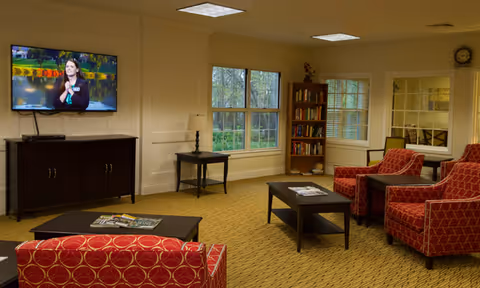 A cozy living room area with red patterned armchairs, a coffee table with magazines, a dark wooden TV stand with a flat-screen TV mounted above it, a side table with a lamp, a bookshelf filled with books, and large windows letting in natural light.