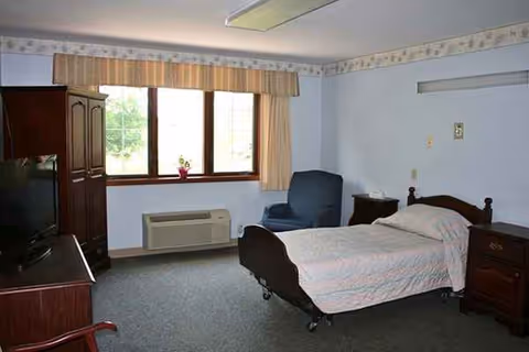 Single bed in a blue-walled senior living room with window, chair, and dresser.