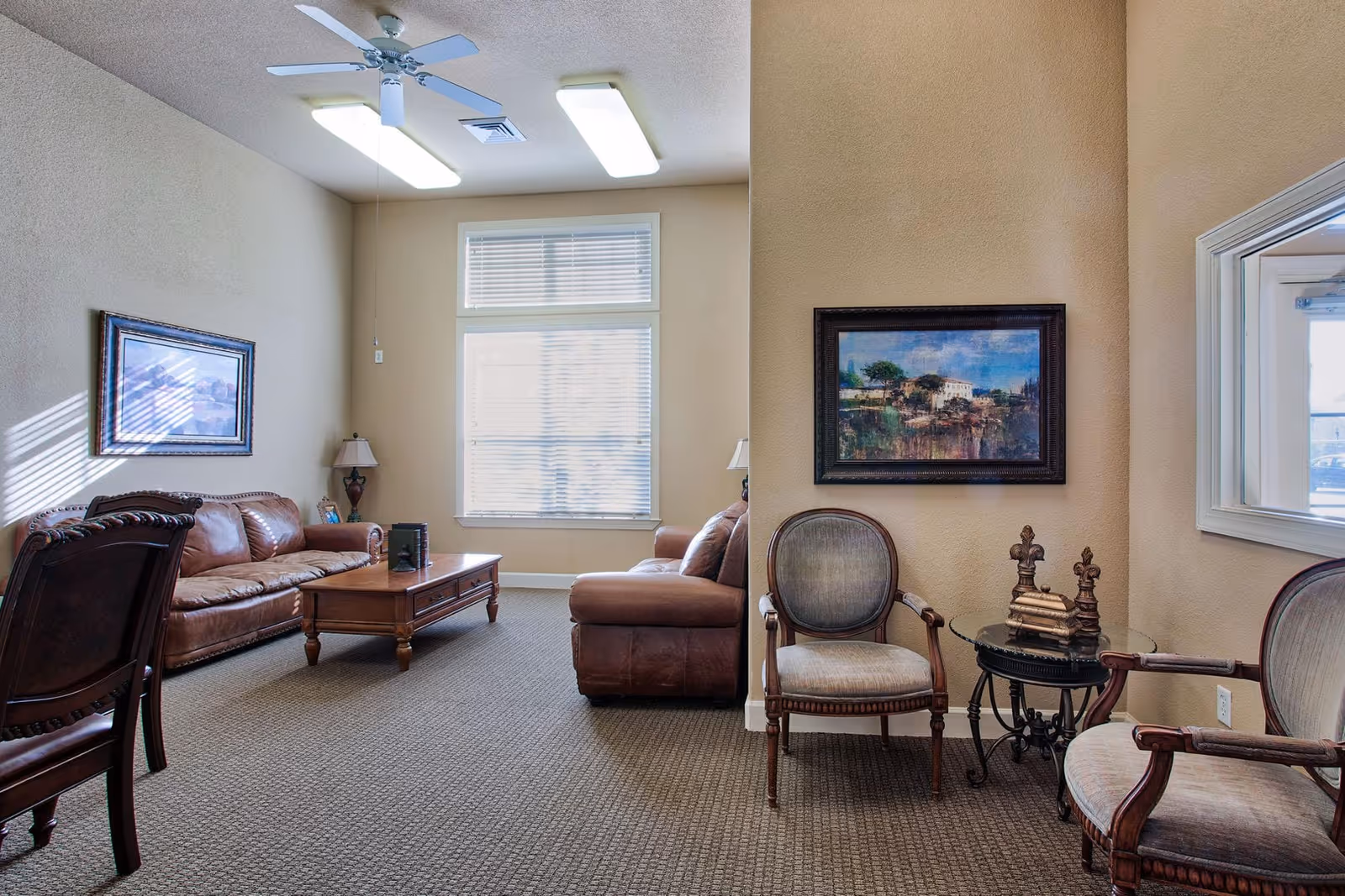 A cozy living room area with beige walls and carpeted floor. The room features two brown leather sofas, a wooden coffee table, two upholstered armchairs with wooden frames, and a small round glass-top side table with decorative items. There are two framed paintings on the walls, a ceiling fan, and fluorescent ceiling lights. A window with blinds allows natural light into the room.