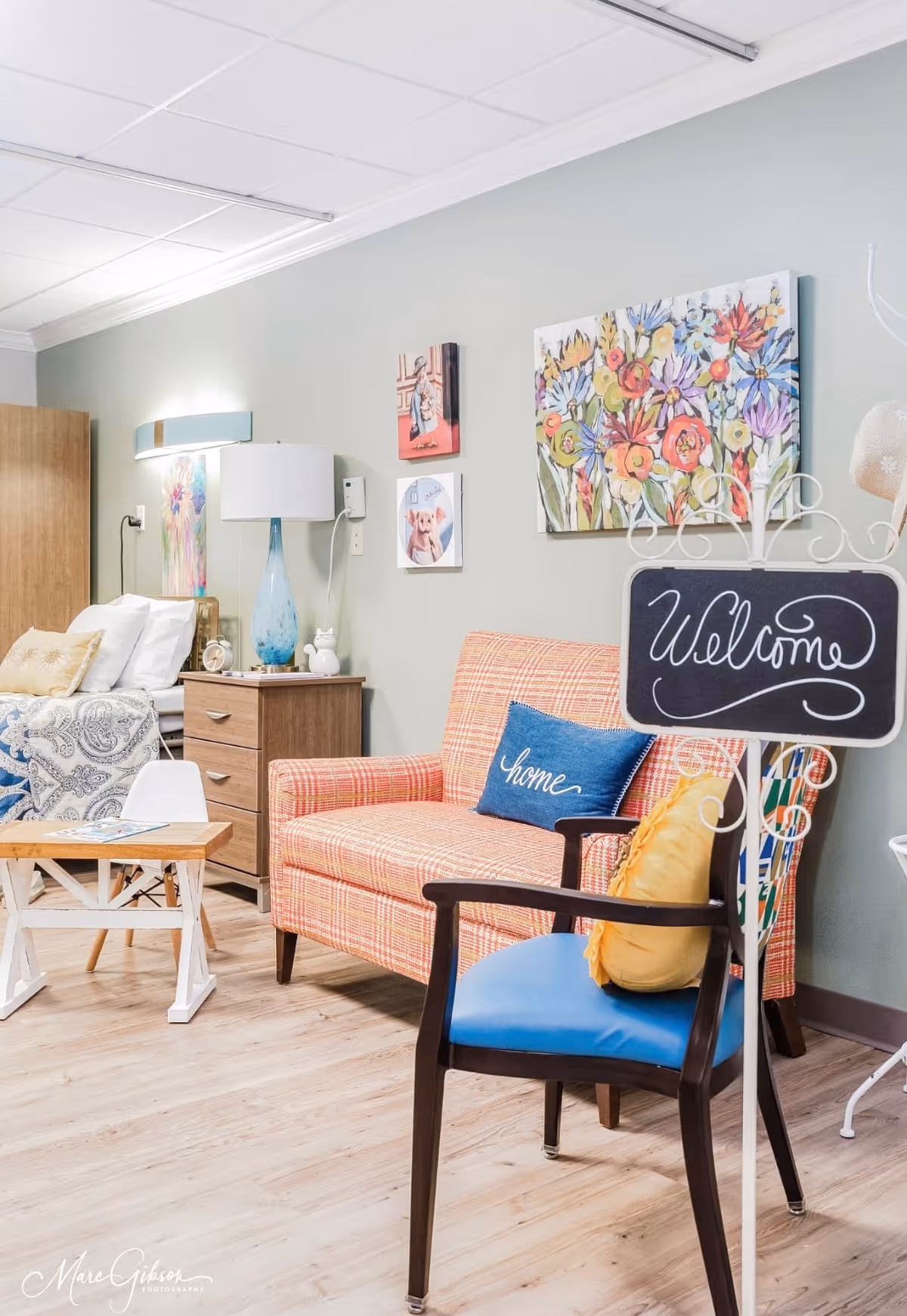 A cozy, well-decorated senior living room area with a plaid loveseat, blue chair, bedside table, bed and floral wall art.