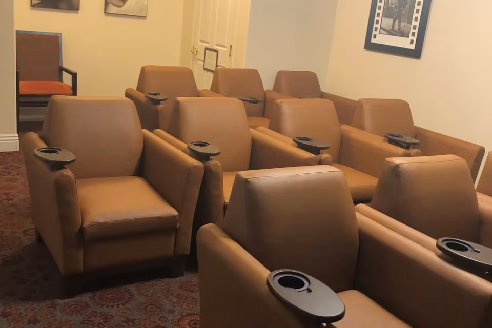 A small theater room with two rows of brown leather recliner chairs, each equipped with a black cup holder on the armrest. The room has a patterned carpet and light-colored walls with framed pictures hanging.