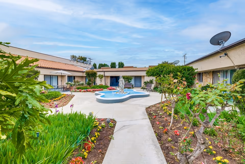 Outdoor courtyard area of a senior living facility with a central water fountain featuring a statue, surrounded by flower beds and greenery. The courtyard is enclosed by single-story buildings with windows and patio furniture, under a partly cloudy sky.