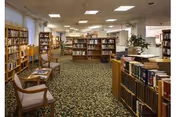 Carpeted library room with bookshelves, chairs, and reading tables under fluorescent ceiling lights.