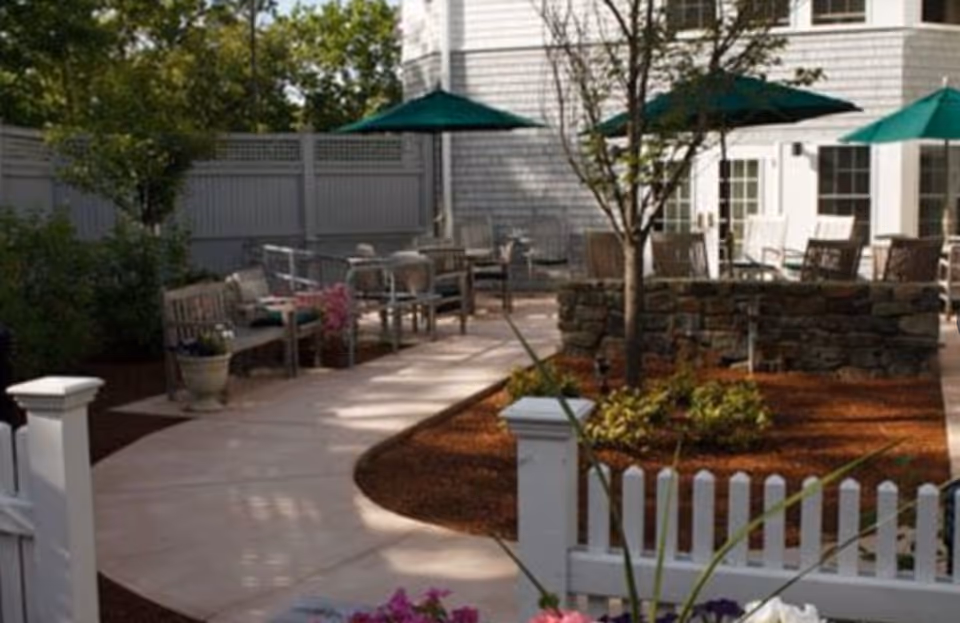 Outdoor patio area with a paved walkway, white picket fence, several wooden benches and chairs, green umbrellas providing shade, trees, and plants surrounding the seating area. The background shows part of a white building with windows and doors.