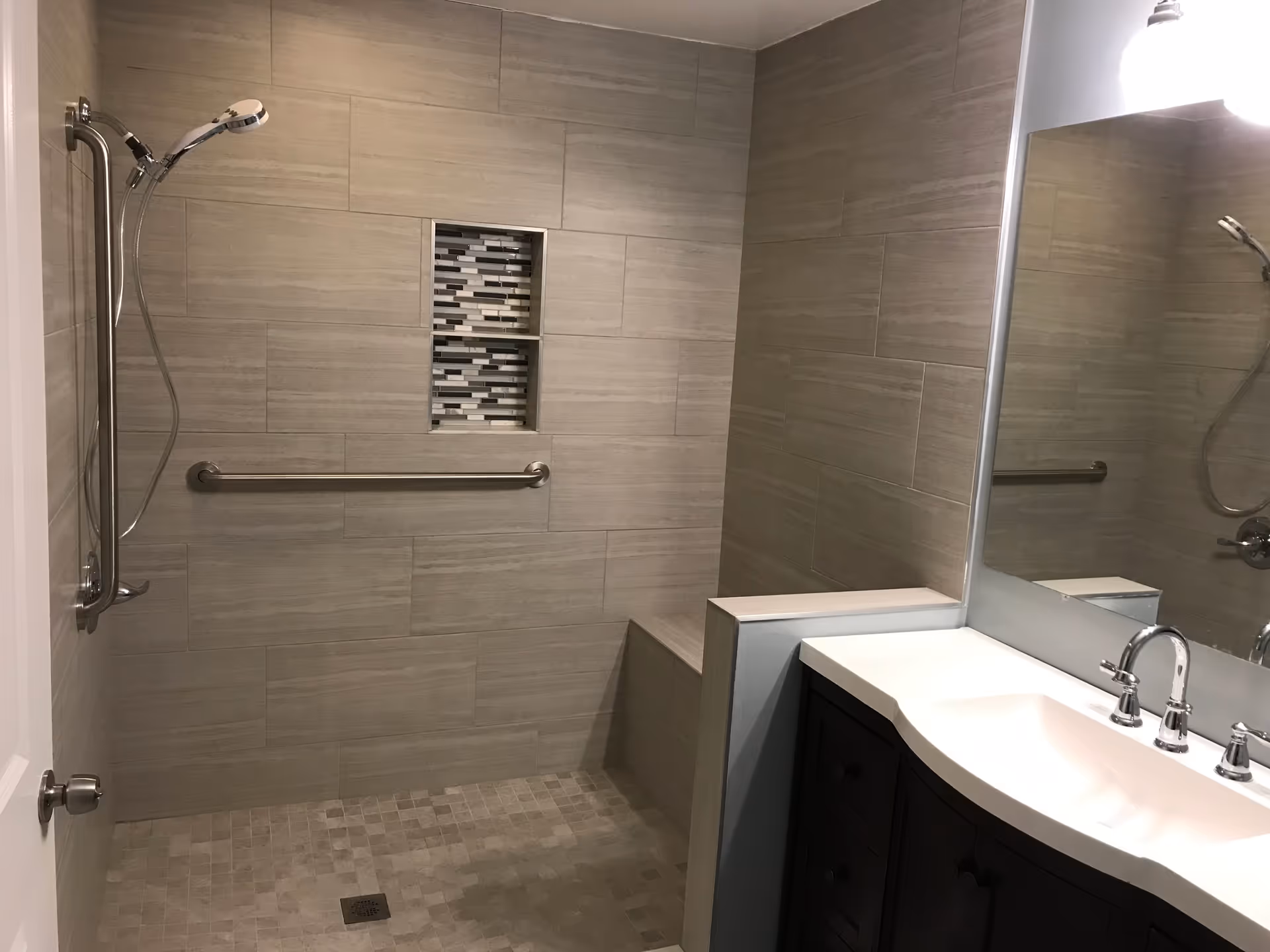 A modern bathroom with a walk-in shower featuring beige tiles, a built-in bench, a handheld showerhead, and a horizontal grab bar. There is a recessed shelf with decorative black, white, and gray tiles. A white sink with a chrome faucet and a large mirror are visible on the right side.