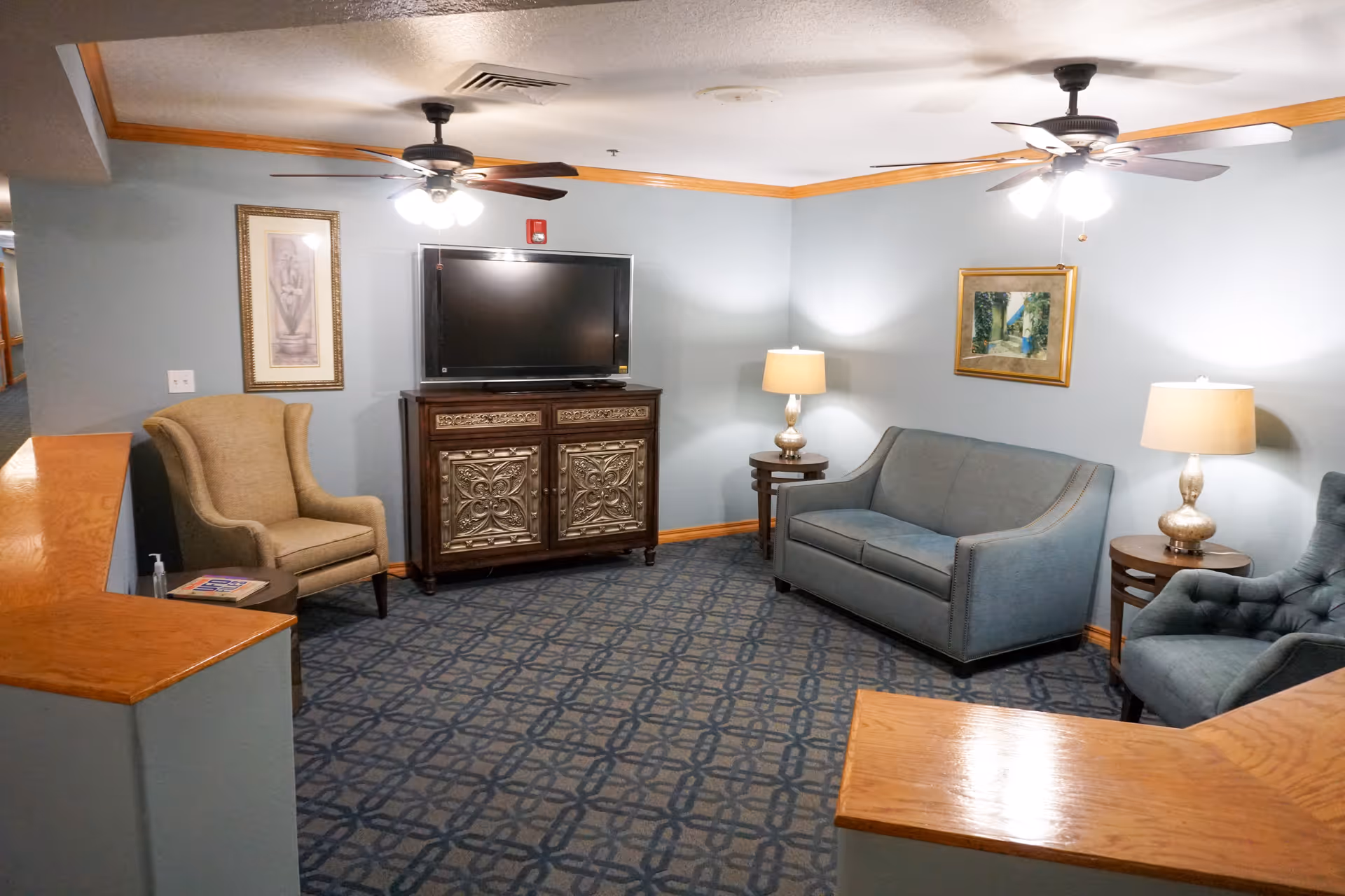 A cozy living room area with a patterned carpet, two ceiling fans with lights, a flat-screen TV on an ornate wooden cabinet, a blue sofa, two blue armchairs, two side tables with lamps, and framed artwork on the light blue walls.