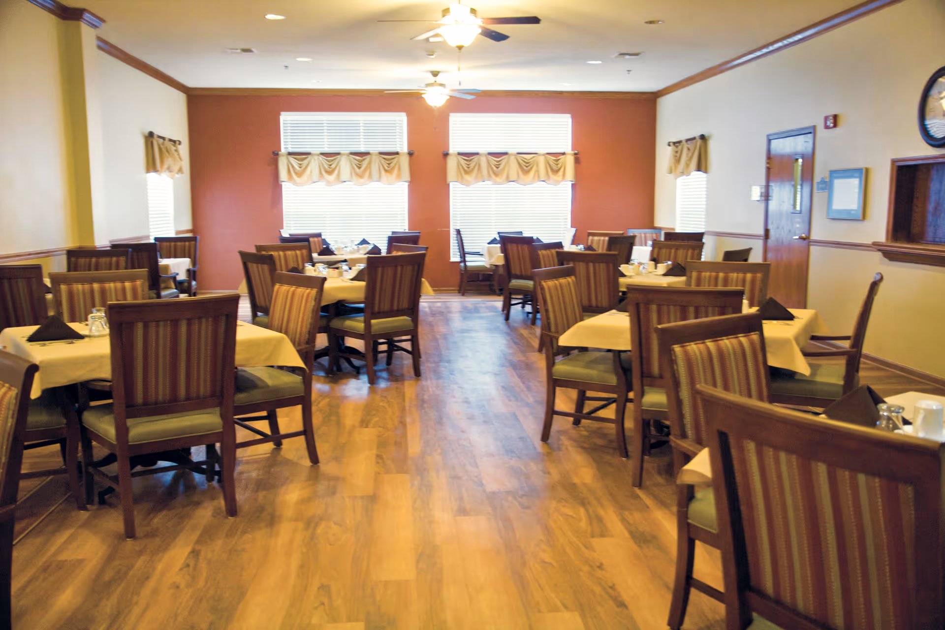 Dining room with multiple tables and wooden chairs set with napkins and tableware and windows at the far wall.