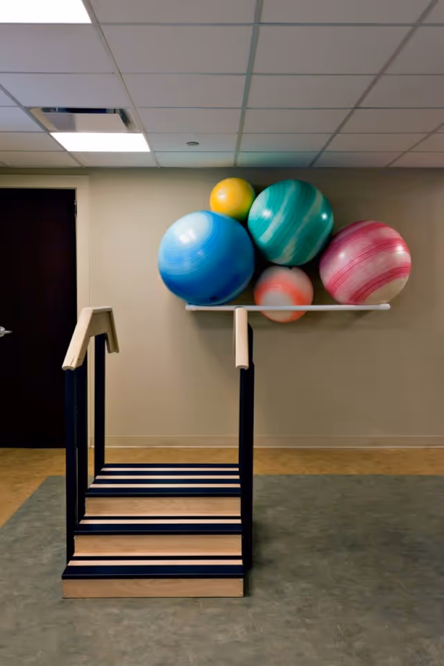 Indoor physical therapy area with a small set of wooden stairs with handrails on both sides and a wall-mounted rack holding five large colorful exercise balls in blue, green, yellow, pink, and red.