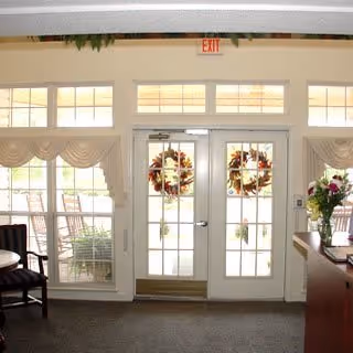 Interior view of a room with double glass doors decorated with autumn wreaths. The doors lead to an outdoor patio area with rocking chairs. There are windows above and beside the doors with beige curtains. A wooden chair and a counter with a vase of flowers are visible inside the room.