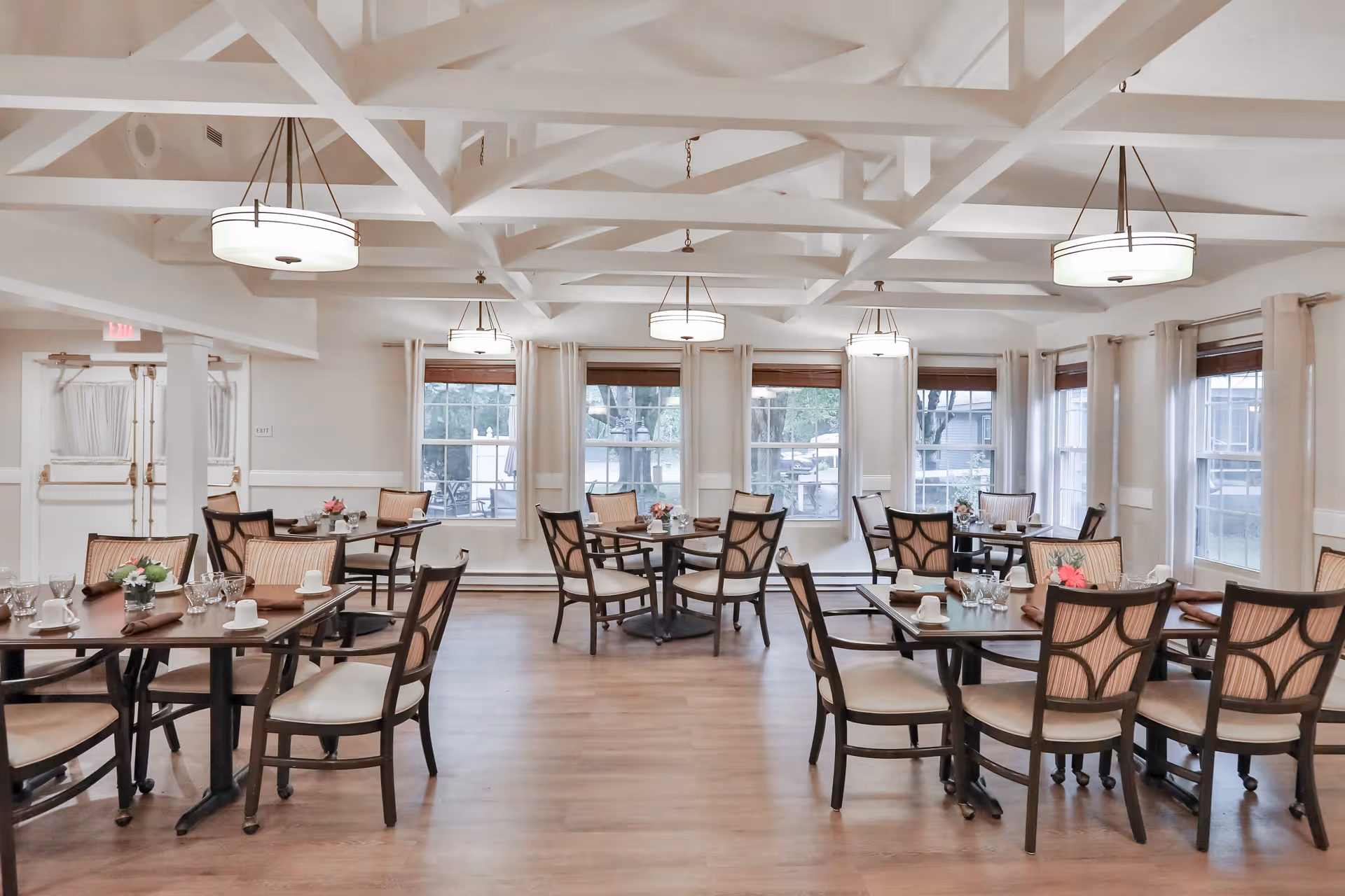 A bright and spacious dining room with multiple wooden tables and cushioned chairs arranged neatly. Each table is set with cups, glasses, napkins, and small floral centerpieces. Large windows with white curtains allow natural light to fill the room, and modern ceiling lights hang from the white exposed beam ceiling.