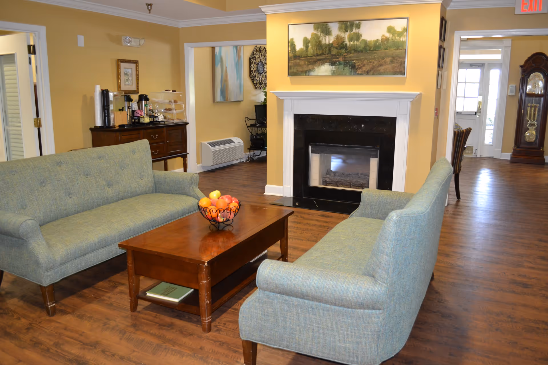 Cozy living room with two green sofas facing a wooden coffee table and a fireplace, artwork on the wall and a fruit bowl on the table.