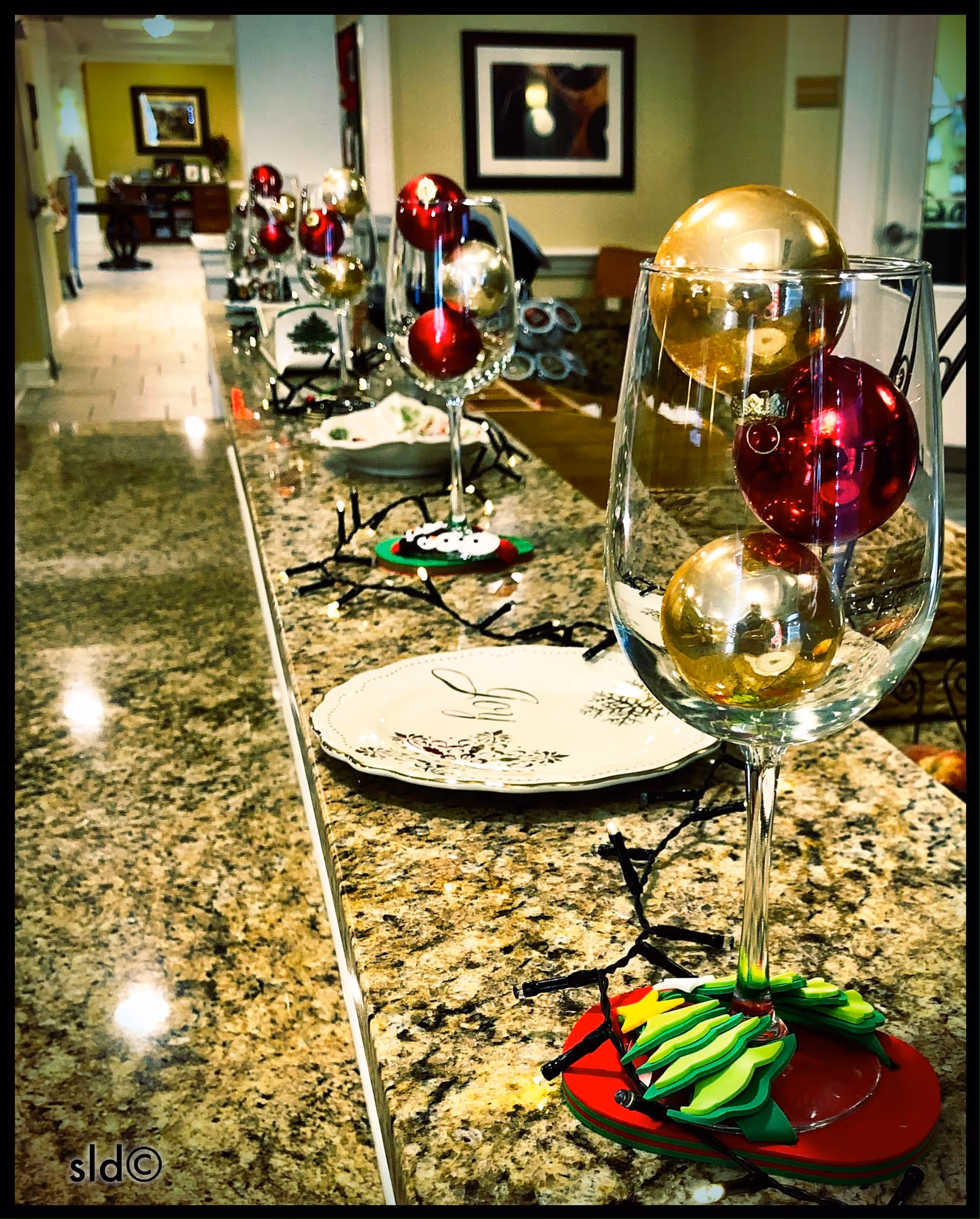 A granite countertop in a dining area lined with wine glasses filled with red and gold Christmas ornaments and place settings.