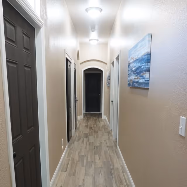 A narrow hallway with beige walls and wood-patterned tile flooring. There are multiple dark brown doors on both sides of the hallway and two ceiling lights illuminating the space. A blue and white abstract painting is hung on the right wall.