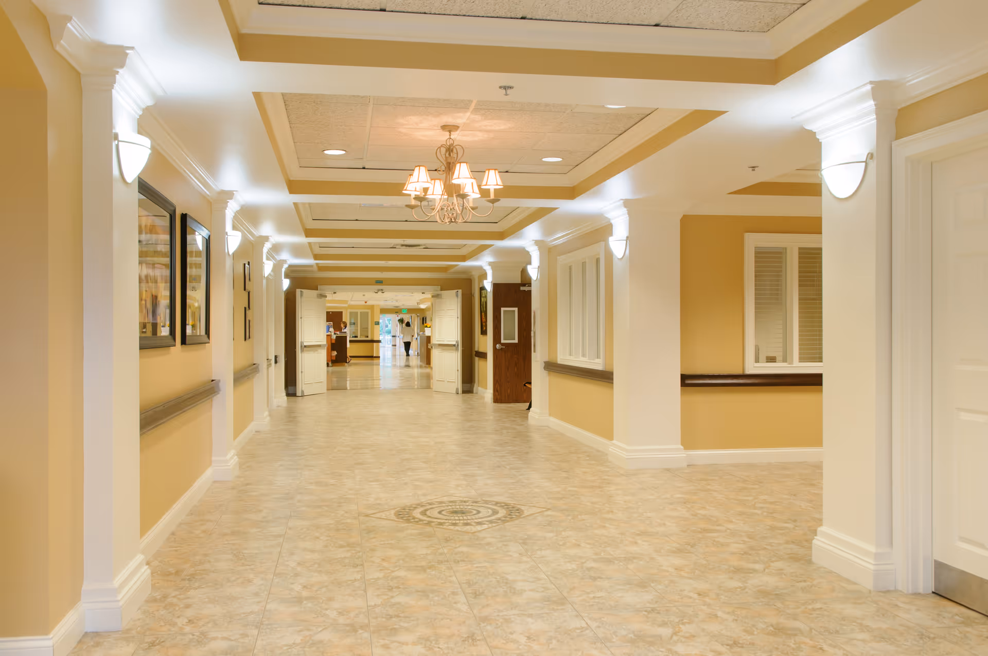 A wide, well-lit hallway in a senior living facility with beige walls, white columns, and tiled floors. The ceiling features recessed lighting and a chandelier. There are framed pictures on the left wall and windows on the right. Double doors are open at the far end of the hallway.