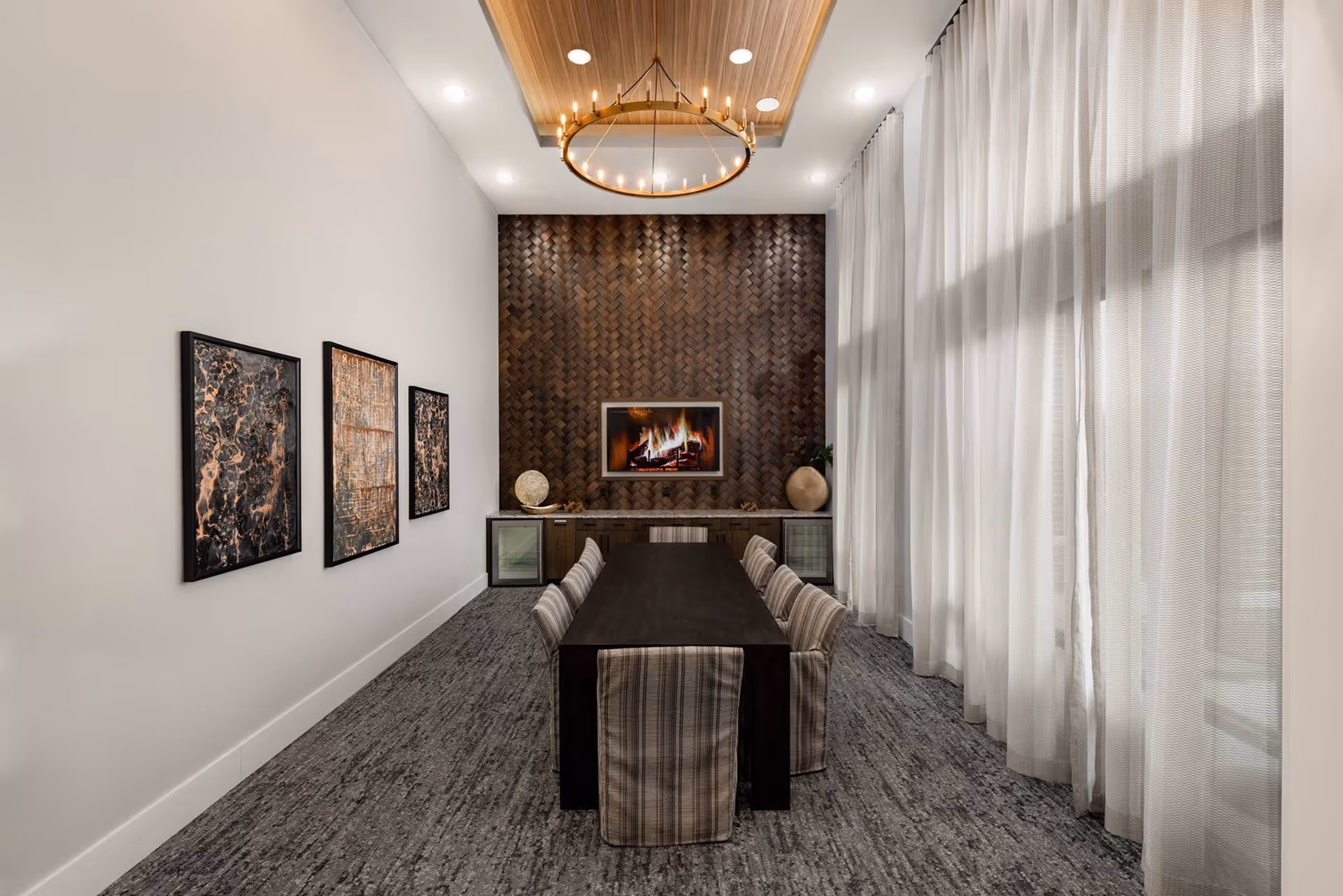 A modern dining room with a long dark wooden table surrounded by eight upholstered chairs with striped fabric. The room features a textured dark wood accent wall with a built-in electric fireplace, flanked by two glass-front cabinets. Three abstract art pieces hang on the left wall, and large sheer white curtains cover the windows on the right. A circular chandelier with multiple small lights hangs from a wooden ceiling panel.