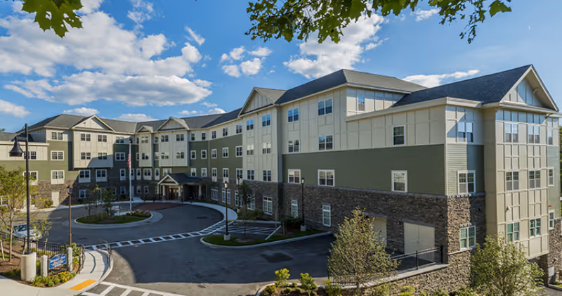 Exterior view of a large, multi-story senior living facility building with a circular driveway, landscaped greenery, and a partly cloudy blue sky.
