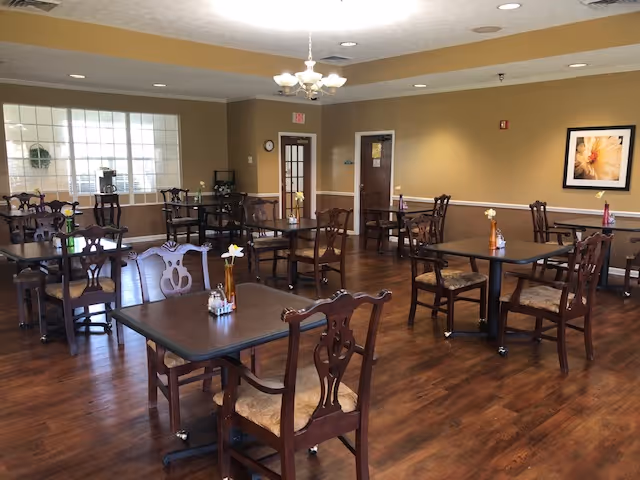 A dining room with several dark wooden tables and matching chairs arranged neatly on a polished wooden floor. Each table has a small vase with flowers and condiments. The walls are painted beige with white trim, and there is a framed flower picture on one wall. A chandelier hangs from the ceiling, and there are windows and doors visible in the background.