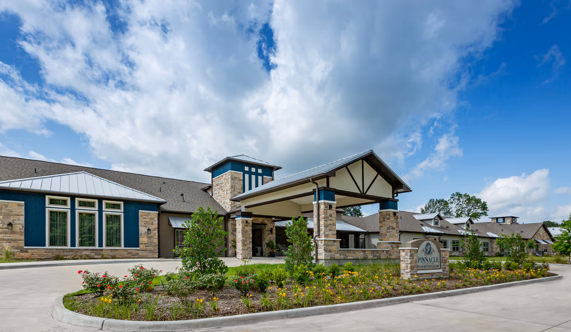 Exterior view of Pinnacle Senior Living facility showing a modern building with stone and blue accents, a covered entrance, landscaped flower beds, and a partly cloudy sky.