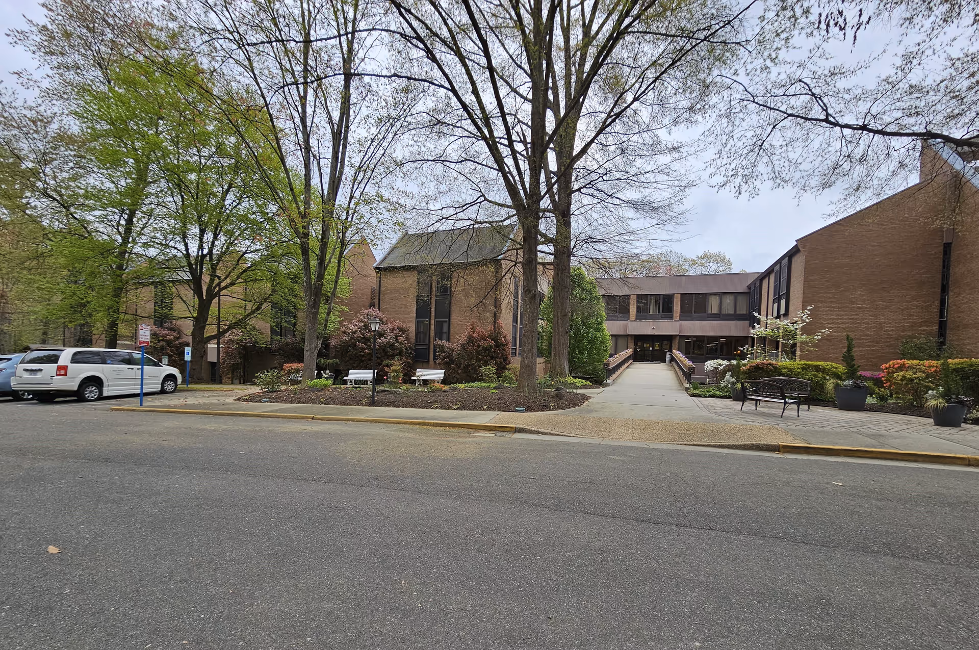 Brick building front with an entrance ramp, benches, trees, and parked cars.