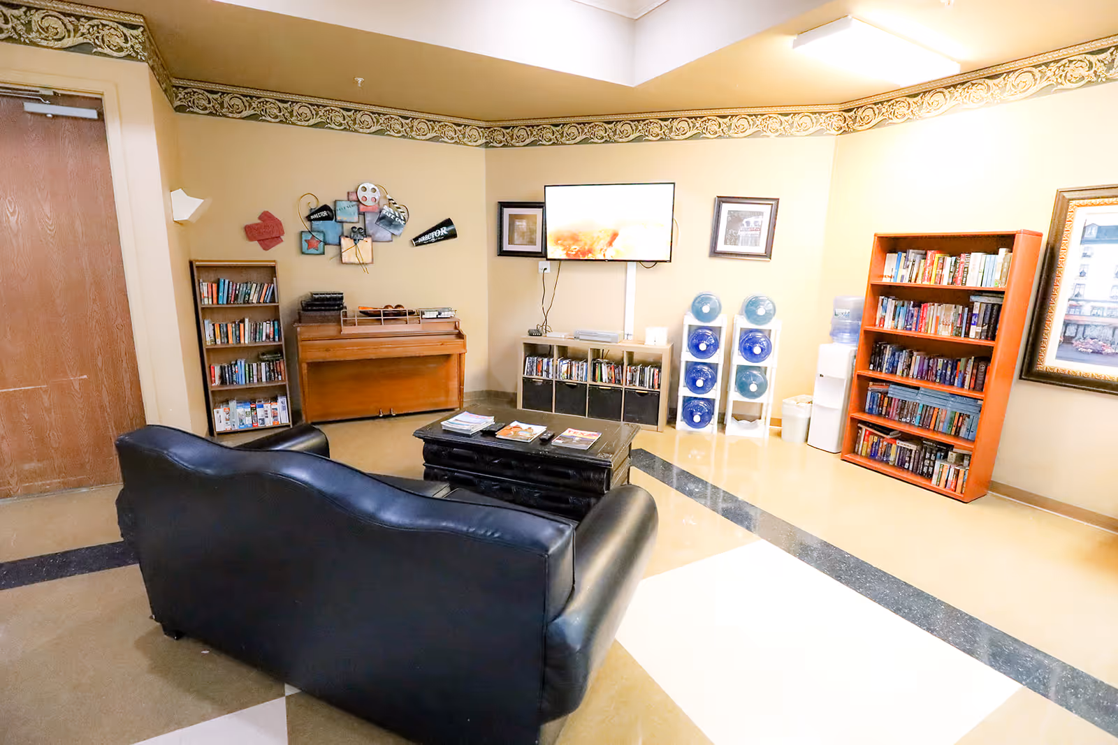 A cozy common area with a black leather couch facing a wall-mounted TV. The room has bookshelves filled with books, a wooden piano, a coffee table with magazines, and water dispensers. The walls are beige with decorative trim near the ceiling.