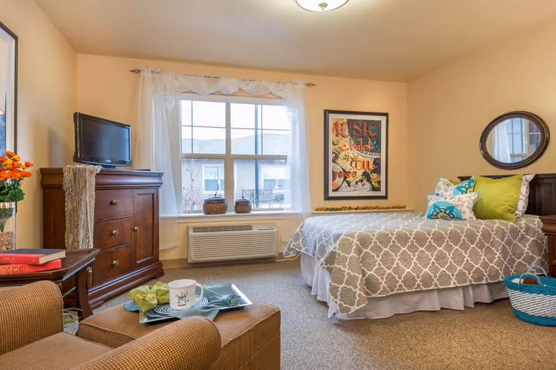 A cozy senior living bedroom with a bed covered in a patterned gray and white bedspread, colorful pillows, a wooden dresser with a small TV on top, a window with sheer white curtains, a framed artwork on the wall, a round mirror, and a comfortable armchair with a tray holding a cup and napkin.